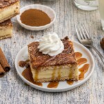 Slice of churro cheesecake served on a small white plate in front of glasses of milk, with a drizzle of caramel sauce and whipped cream on top.