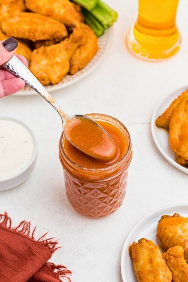 Orange sauce in a small jar, being spooned out, in front of plates of chicken wings.