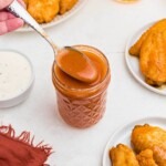 Orange sauce in a small jar, being spooned out, in front of plates of chicken wings.