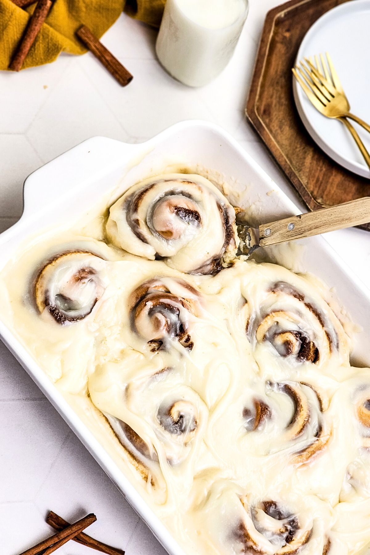 Gooey cinnamon rolls in a white baking dish, topped with icing, and surrounded by plates and glass of milk.