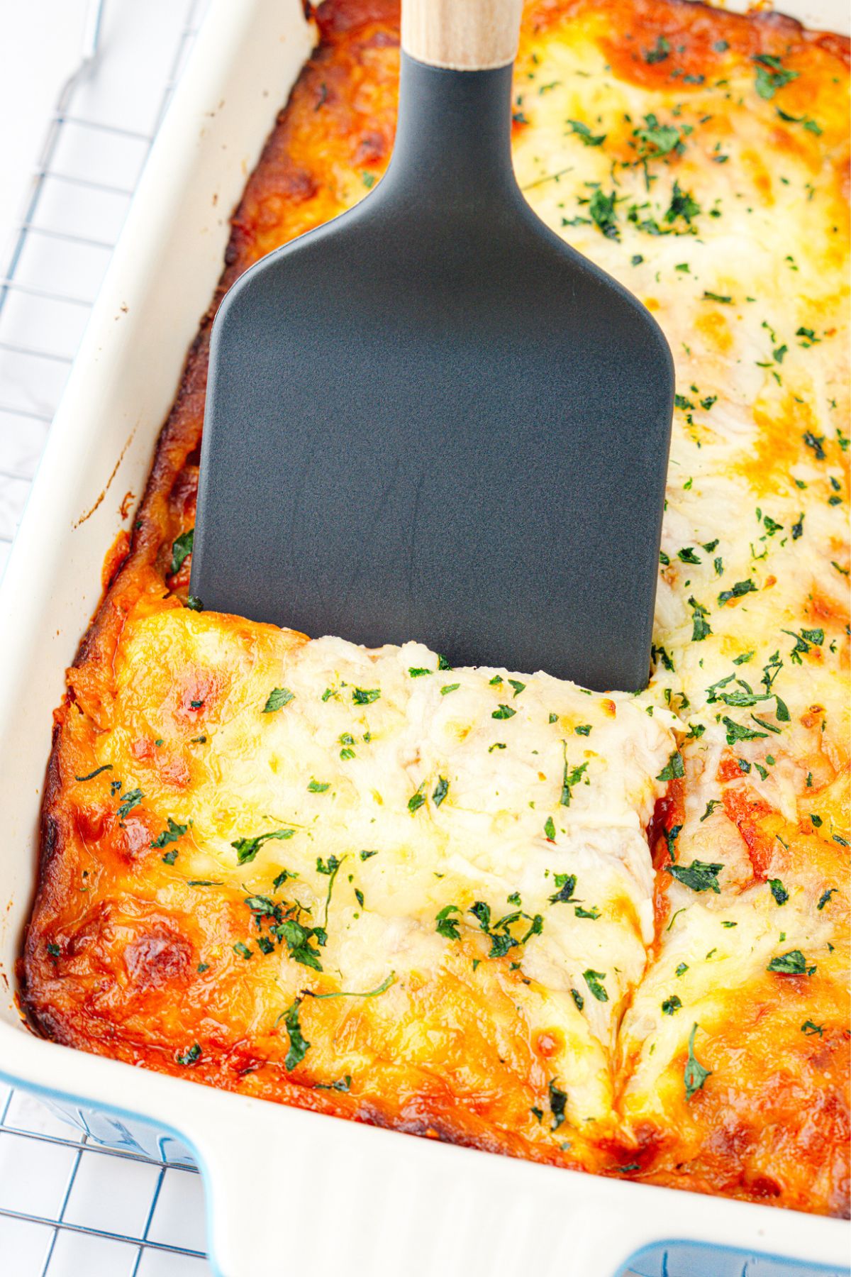 White casserole dish of ravioli lasagna with a slice being scooped out with a spatula. 