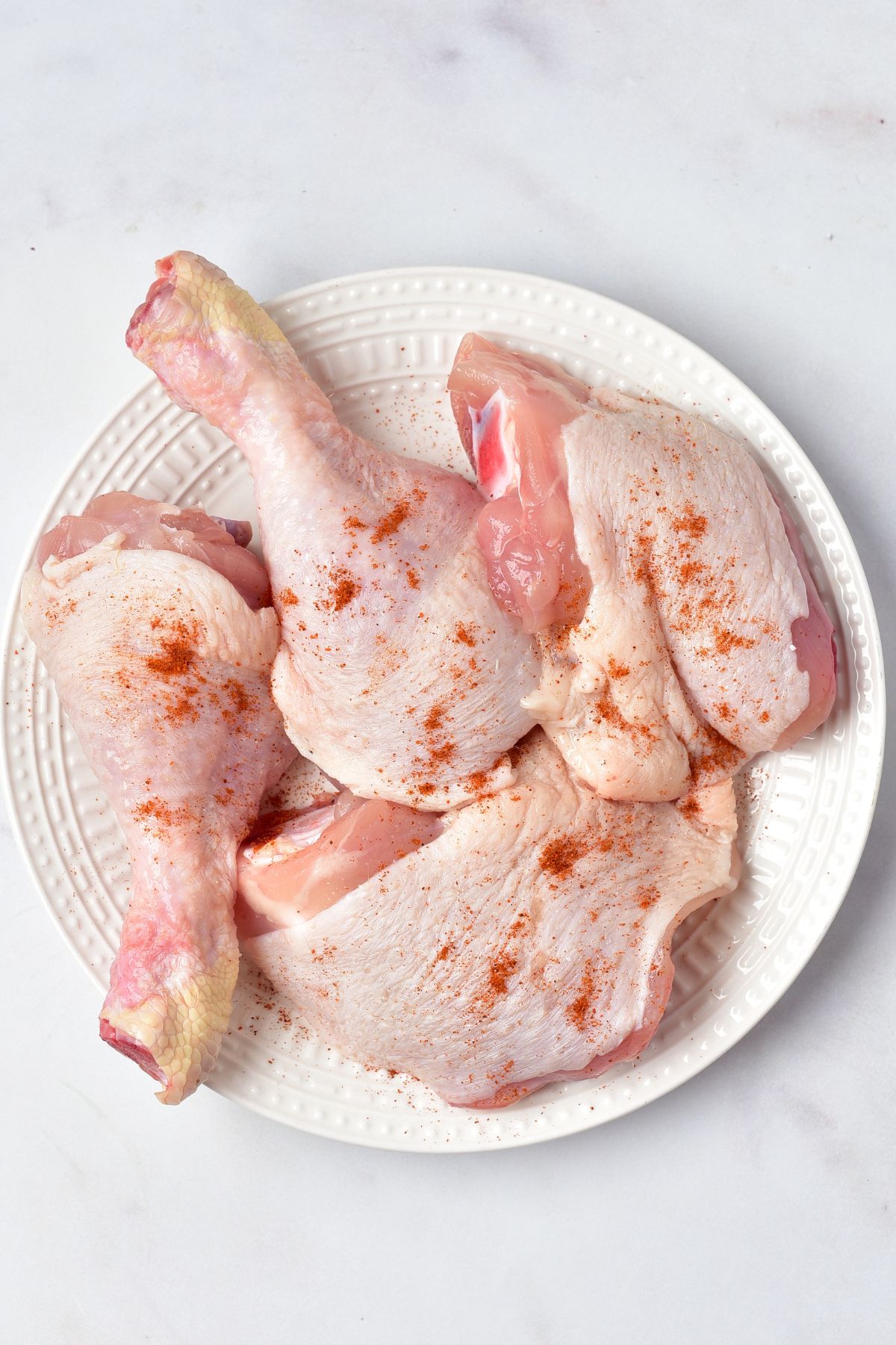 Uncooked chicken, seasoned, on a white plate, on a marble table. 