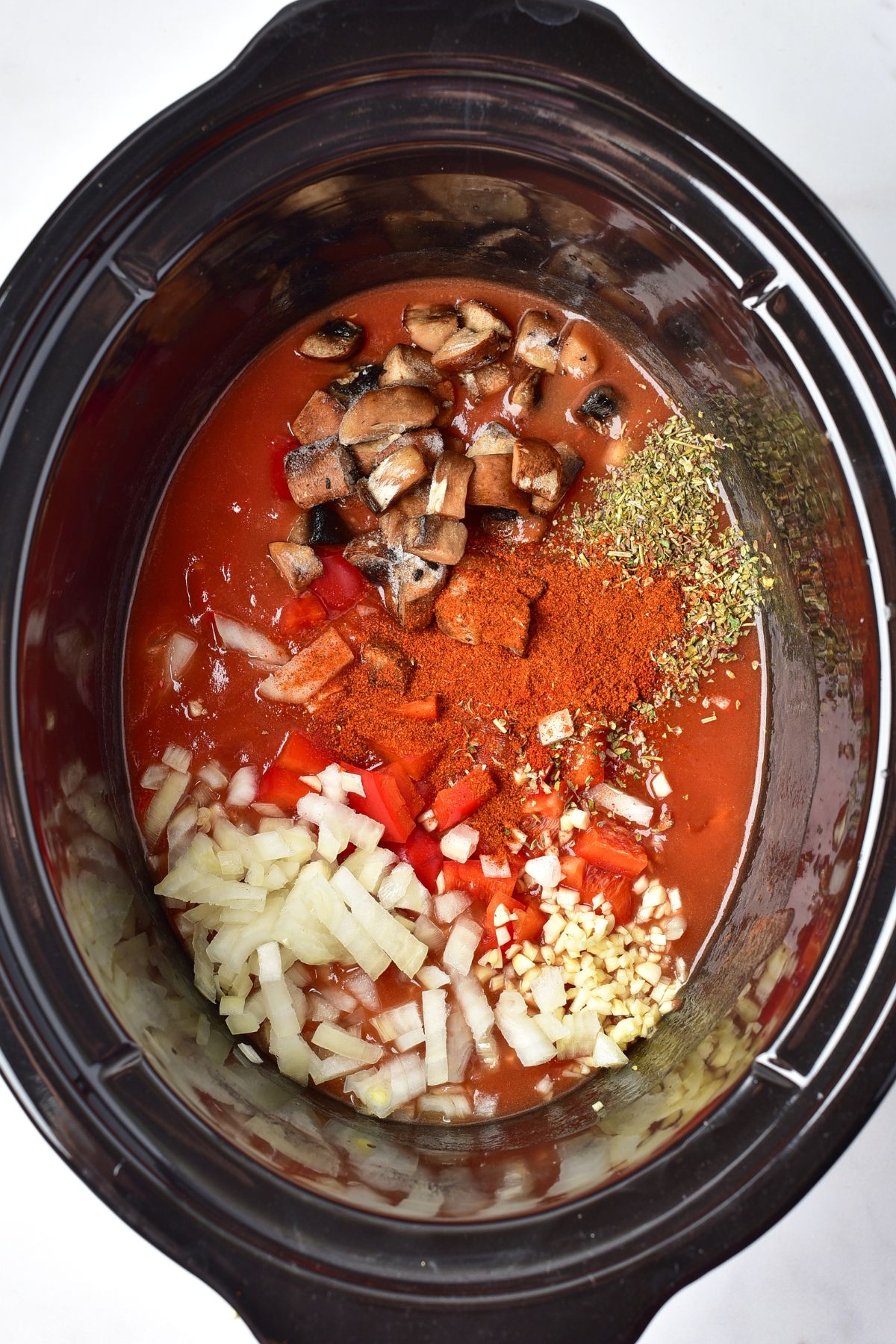 Seasonings, vegetables, and sauce, added to the slow cooker, before being mixed together. 