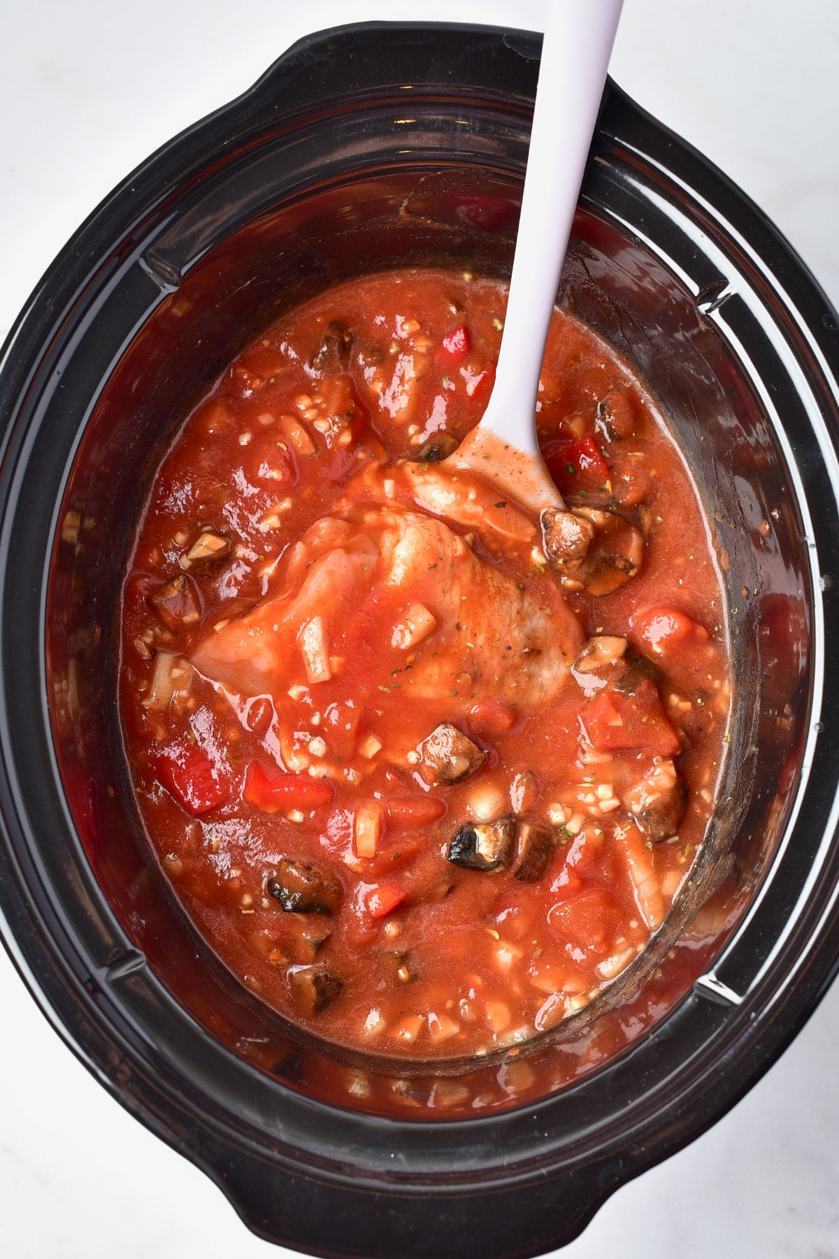 Uncooked chicken added to a slow cooker with seasonings and sauces and vegetables. 