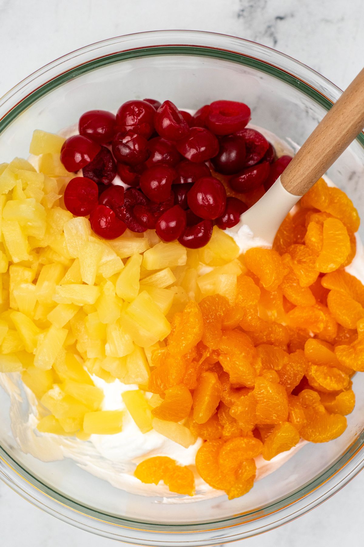 Cherries, pineapple, and mandarin oranges, being added to whipped cream mixture, in a clear glass bowl. 