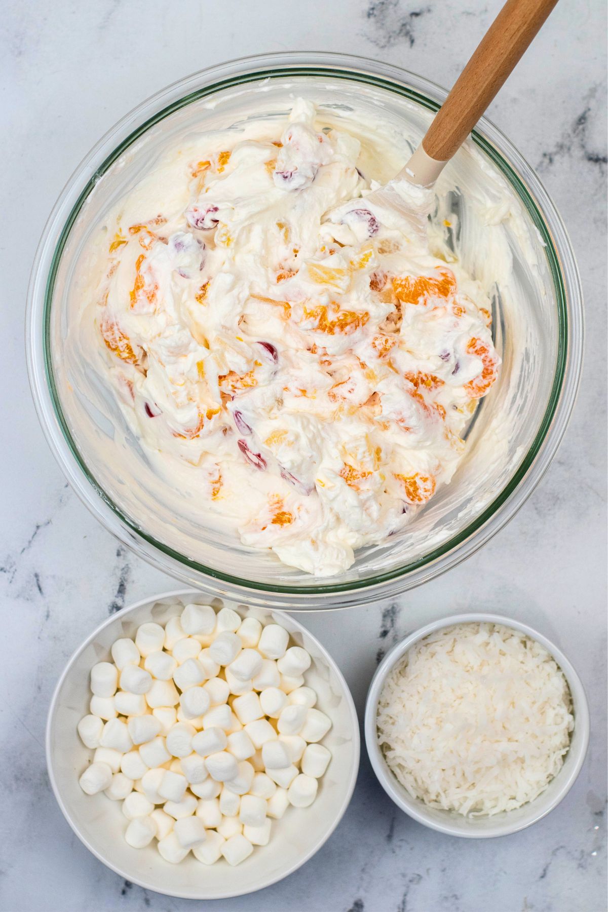 A large clear glass mixing bowl, filled with whipped cream mixture and fruit, having mini marshmallows and coconut. 