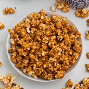 Golden caramel popcorn in a white bowl with pieces of popcorn scattered around the bowl.