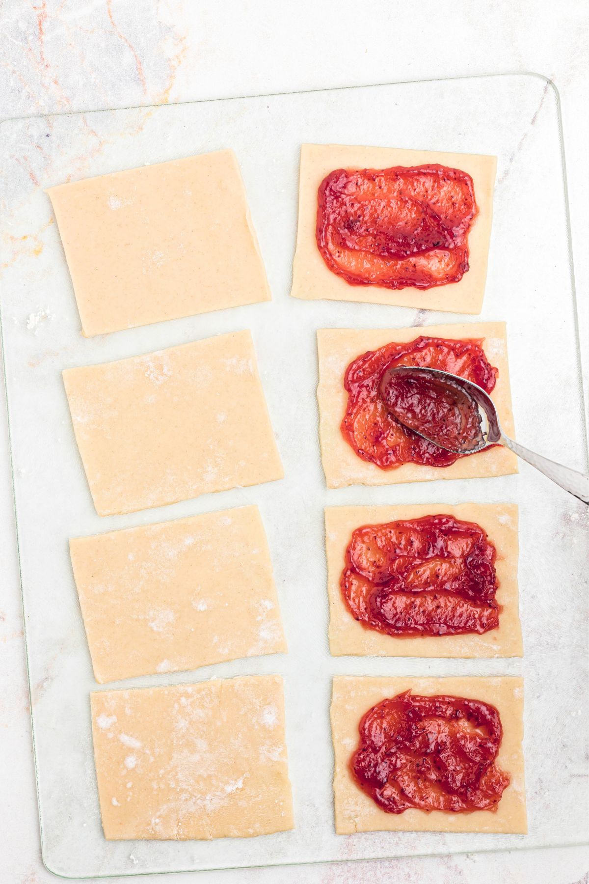 Jam being added by the spoonful to pie crust dough that has been cut into squares. 