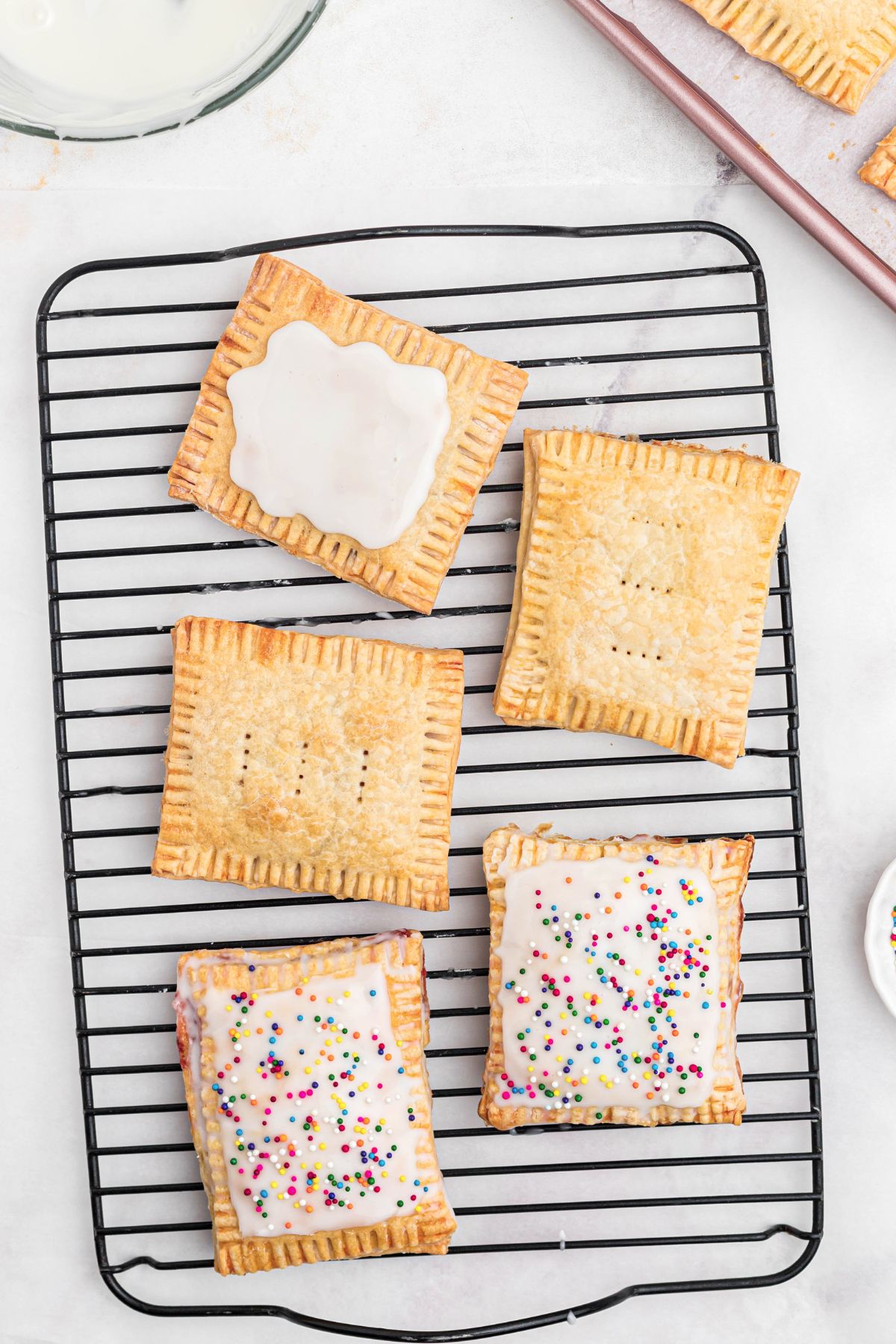 Baked pastries being topped with white icing and sprinkles, on a cooling rack. 