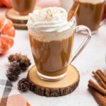 Cup of coffee topped with whipped cream and a cinnamon stick, in front of white pot, on a marble board.