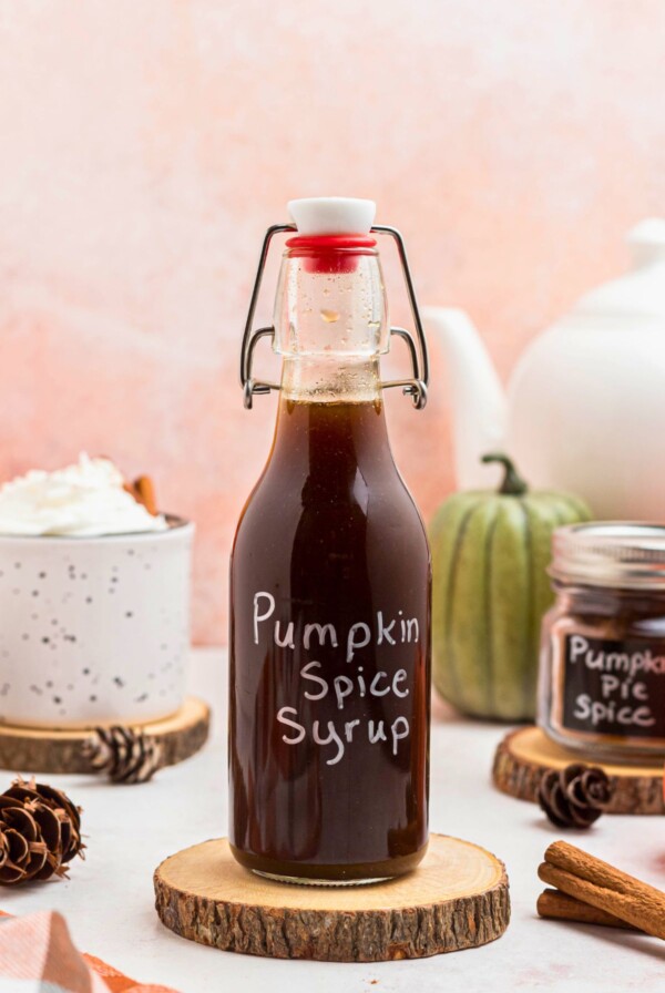 Bottle of pumpkin spice syrup on a small wooden coaster next to a cup of coffee, on a marble table.