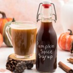 Bottle of pumpkin spice syrup on a small wooden coaster next to a cup of coffee, on a marble table next to a cup of coffee.