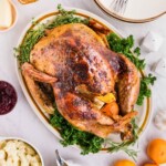 Plated roasted turkey on a serving platter, surrounded by plates, honey, and other food, on a white marble table.