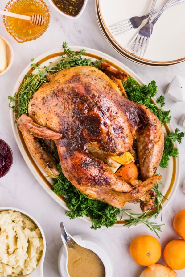 Plated roasted turkey on a serving platter, surrounded by plates, honey, and other food, on a white marble table.