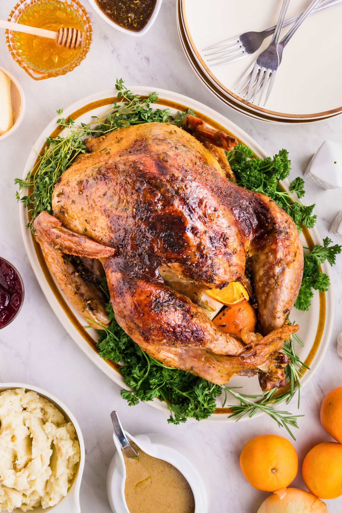 Plated roasted turkey on a serving platter, surrounded by plates, honey, and other food, on a white marble table. 