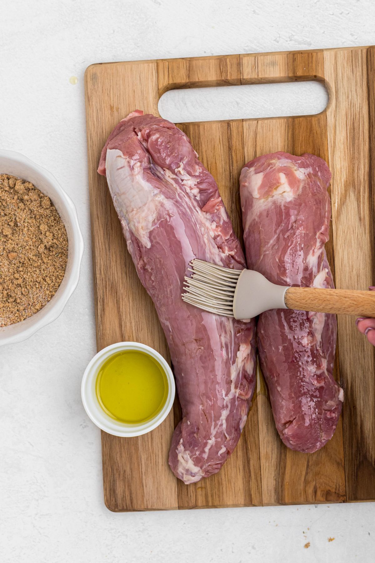 Two uncooked pork tenderloins on a wooden cutting board with olive oil being brushed on top. 