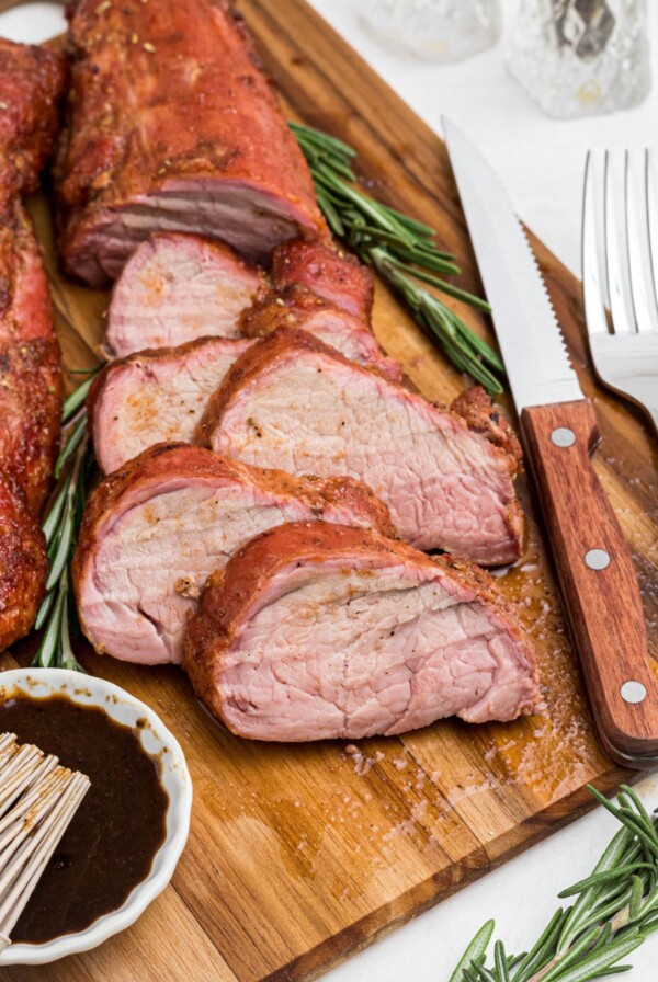 Juicy tenderloin cut into slices on a cutting board with a knife and fork, and fresh rosemary.