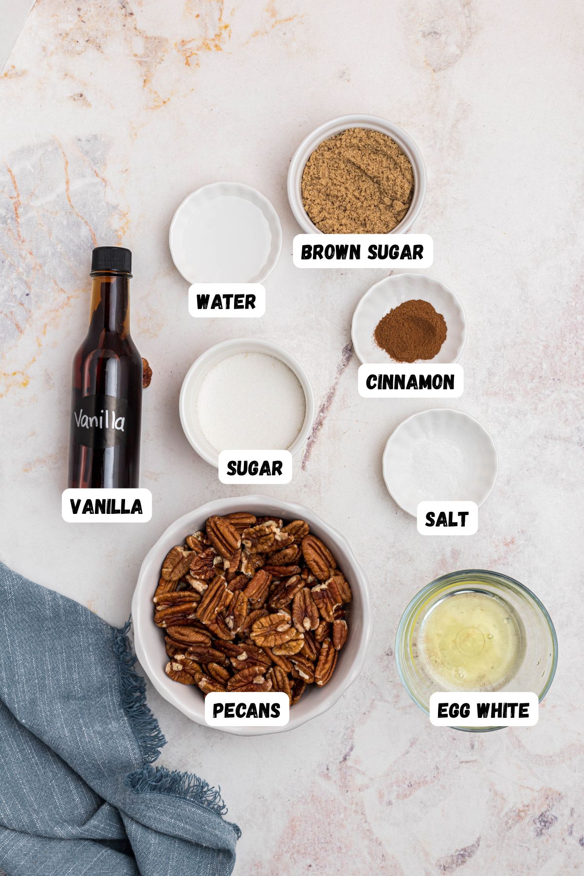 Ingredients needed to make candied pecans measured into small bowls and labeled on a marble table. 