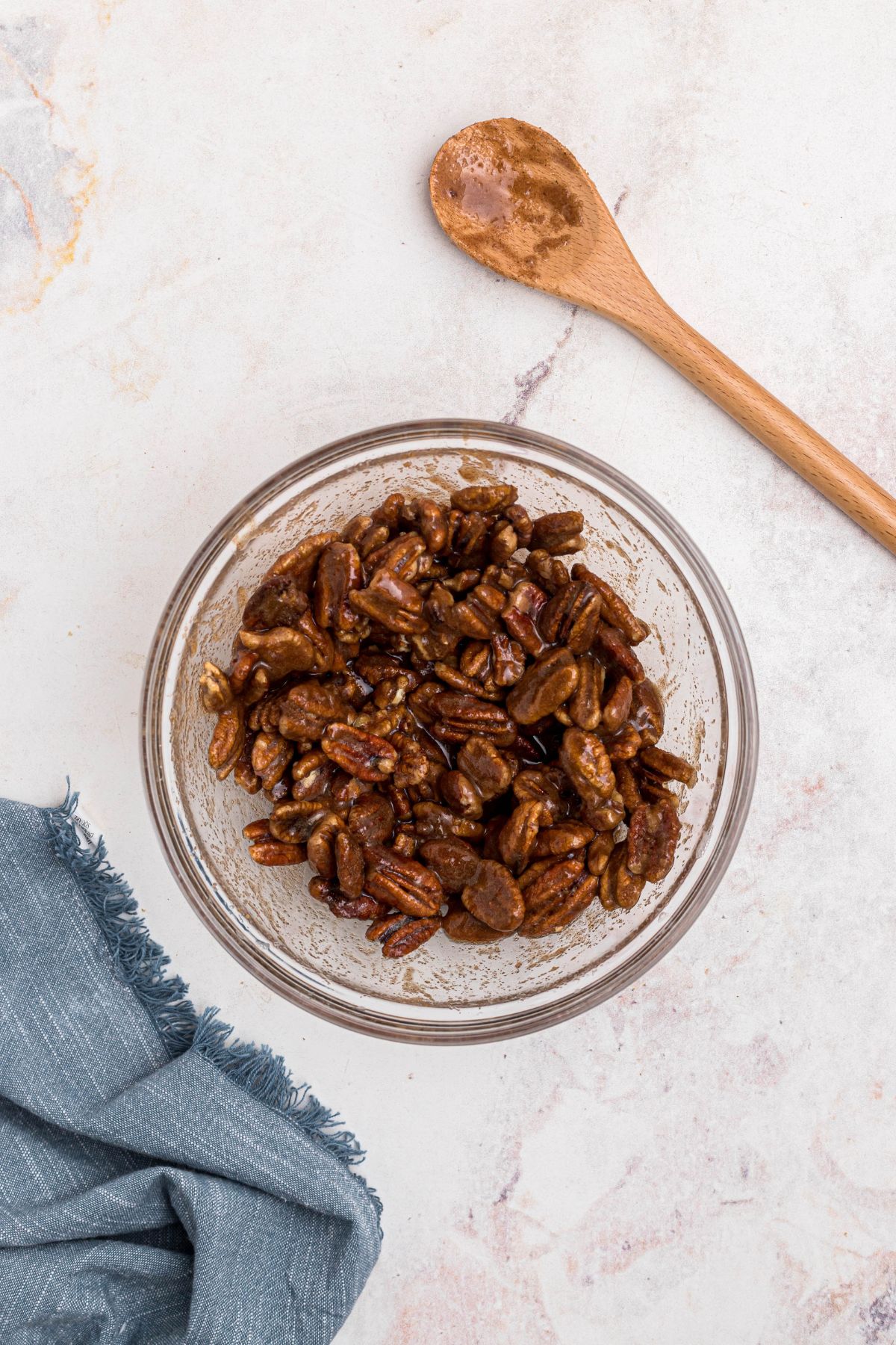 Sugars and cinnamon being tossed with coated pecans in a clear glass bowl on a marble table.