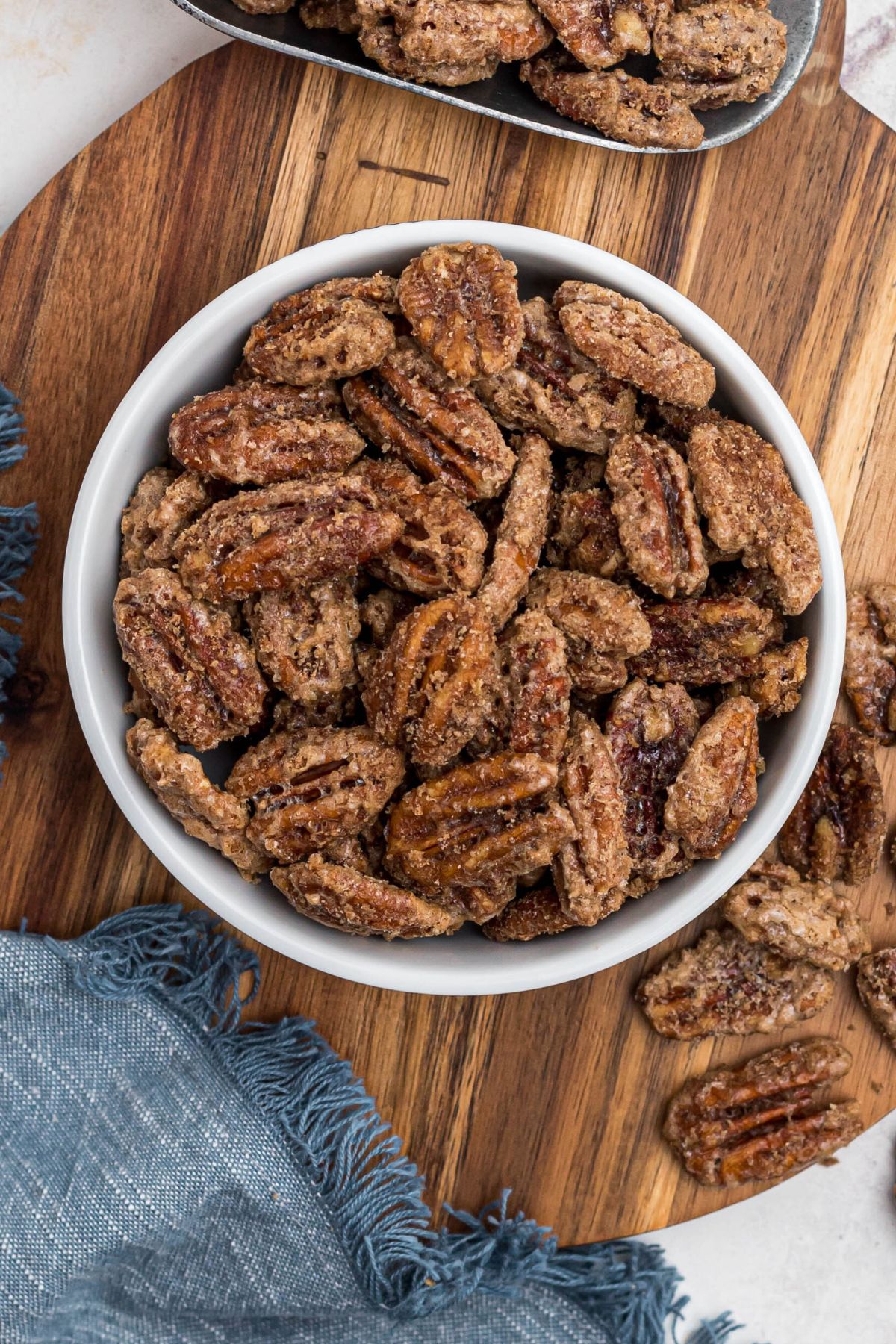 Candied pecans in a small white bowl, with a blue linen and scattered pecans nearby. 