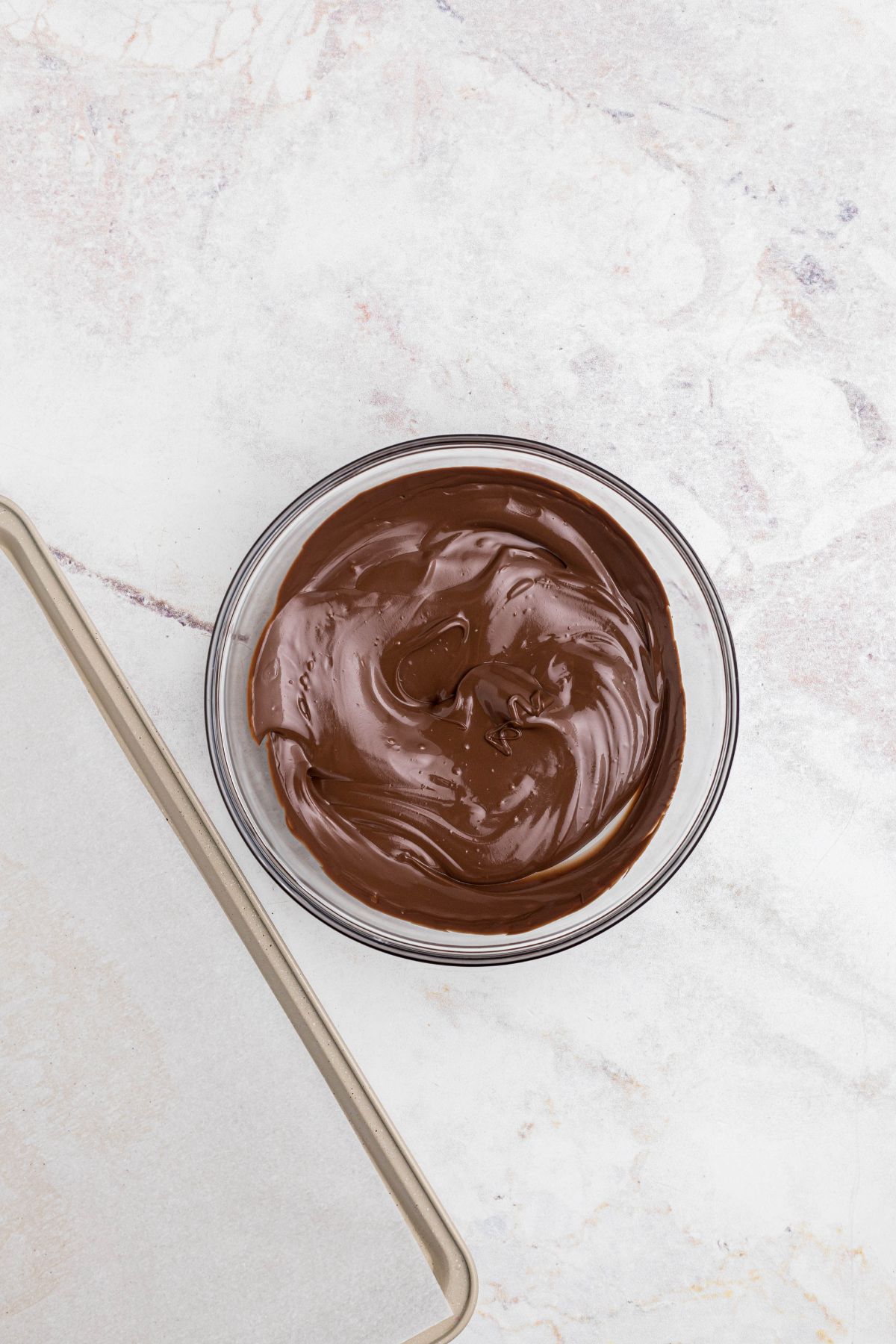 Chocolate melted in a clear glass bowl, next to a baking sheet lined with parchment paper. 
