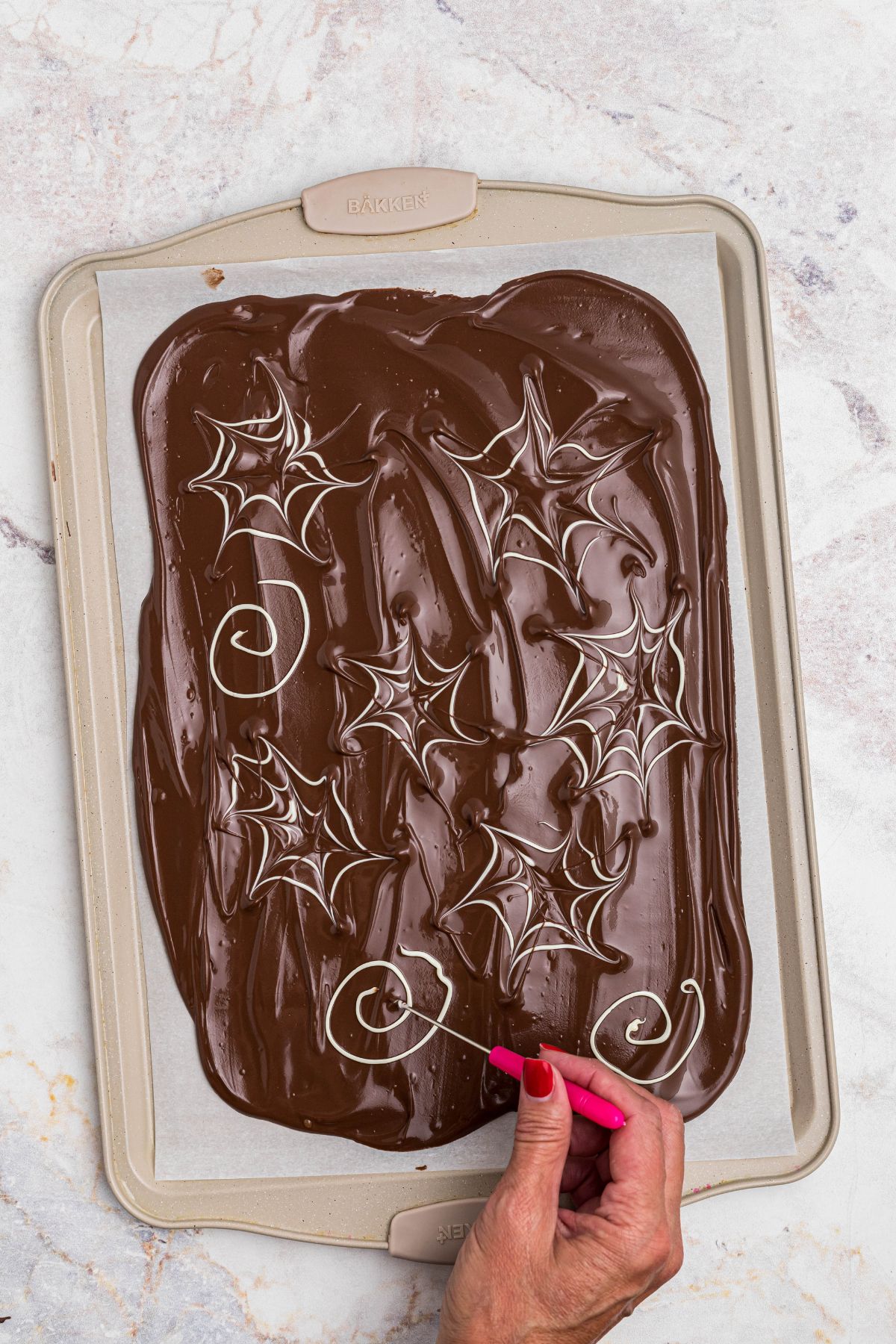 Melted chocolate in a baking sheet, with swirls of melted white chocolate being added, then dragged with a pick tool to make spider web shapes. 