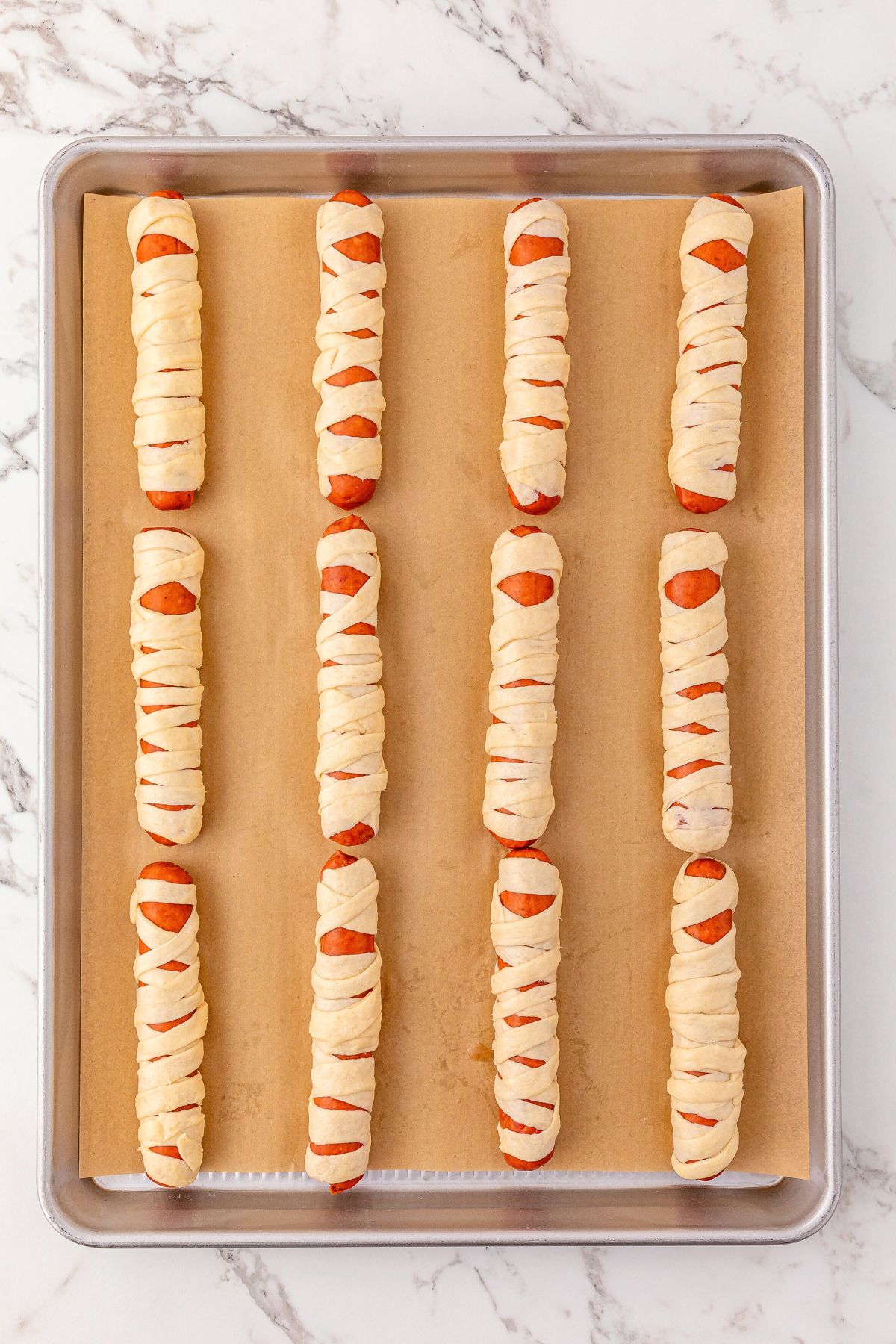 Uncooked hot dogs wrapped with strips of crescent dough, placed on a baking sheet lined with parchment paper.