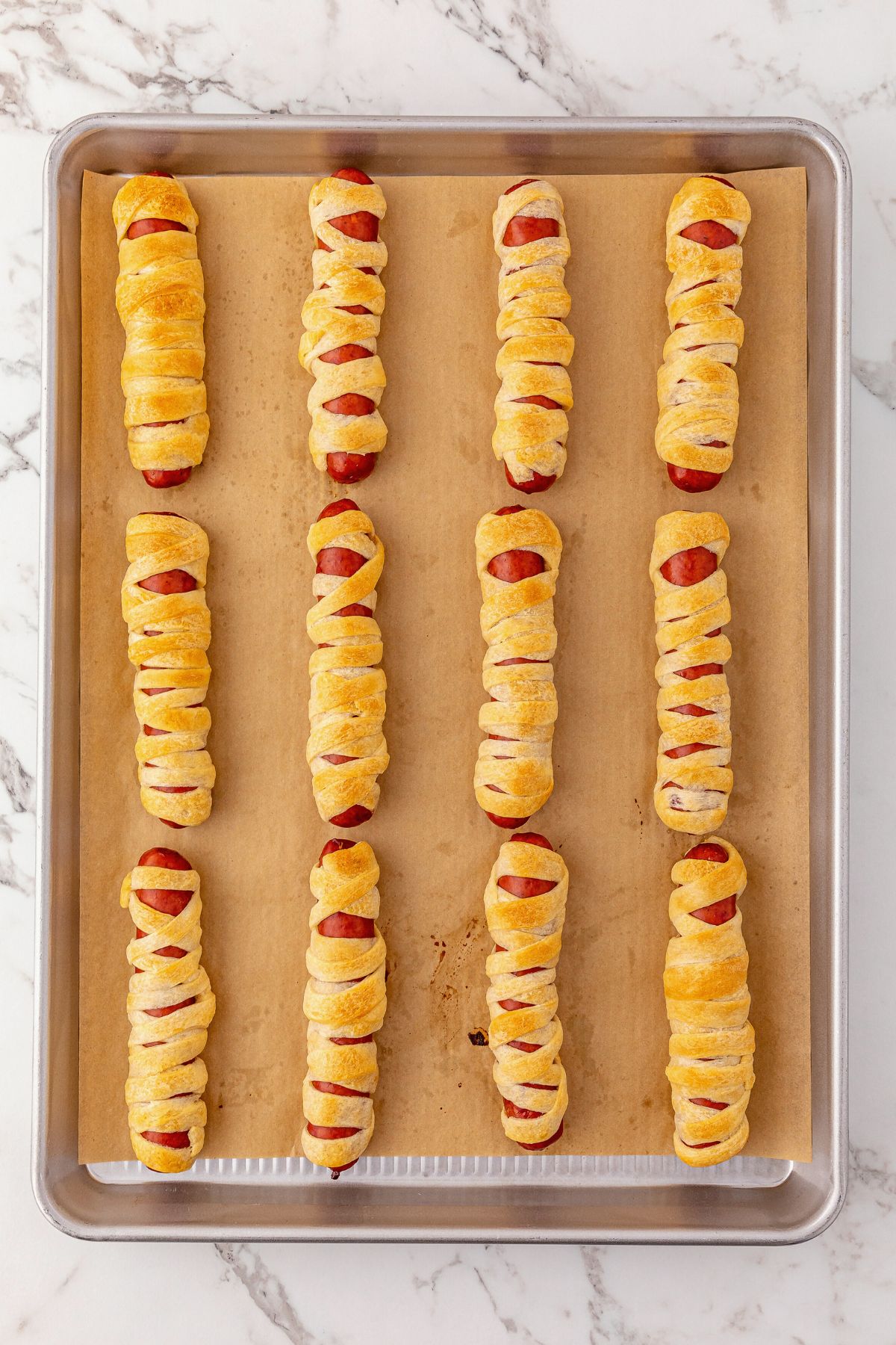 Hot dogs wrapped in strips of dough and baked until golden brown on a baking sheet, lined with parchment paper.