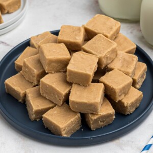 Creamy peanut butter fudge, cut into small pieces, stacked on a blue plate, in front of jars of milk.