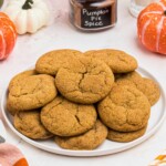 Golden brown pumpkin spice cookies stacked on a white plate in front of glasses of milk.