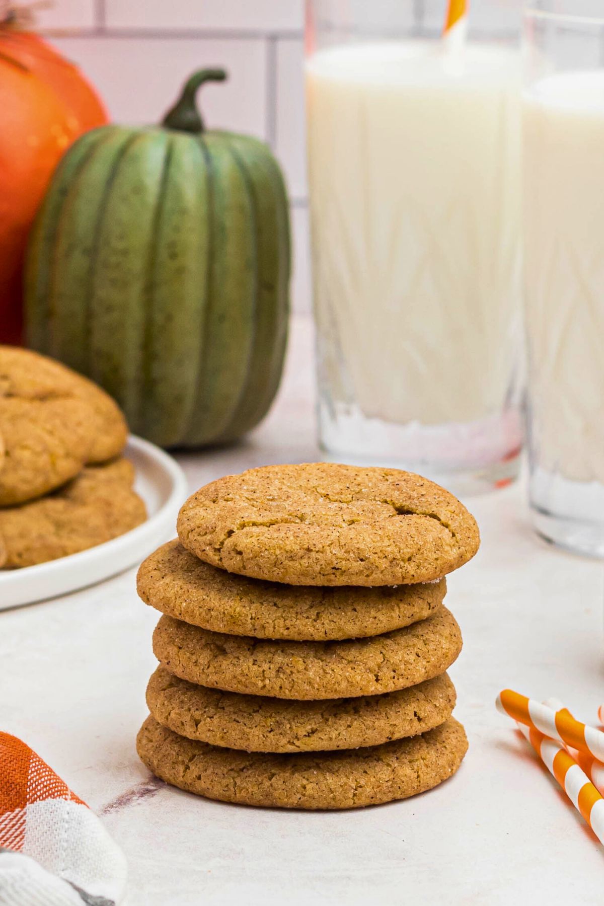 Spice cookies stacked together in front of a large plate of cookies and glasses of milk.