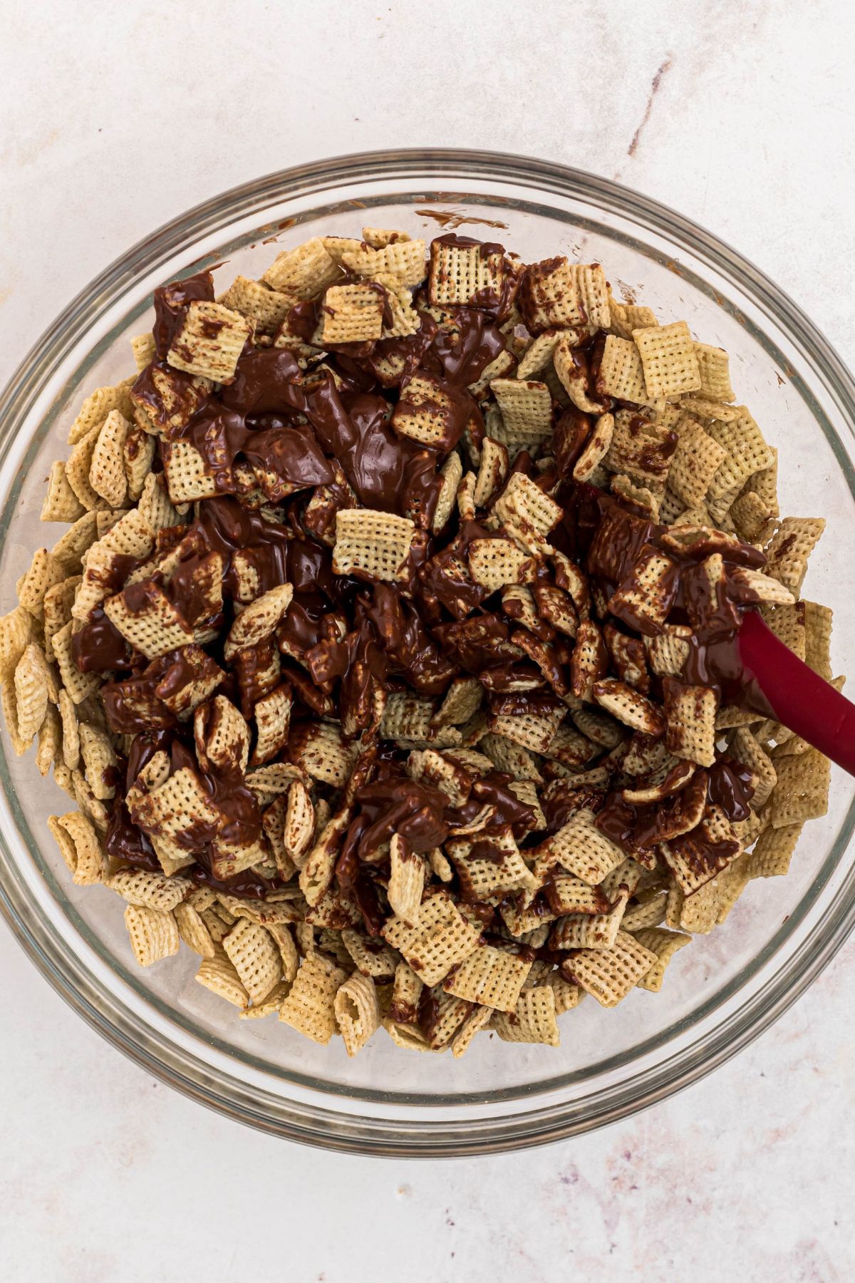 Chocolate and peanut butter mixture being mixed with Chex cereal in a large clear glass bowl. 