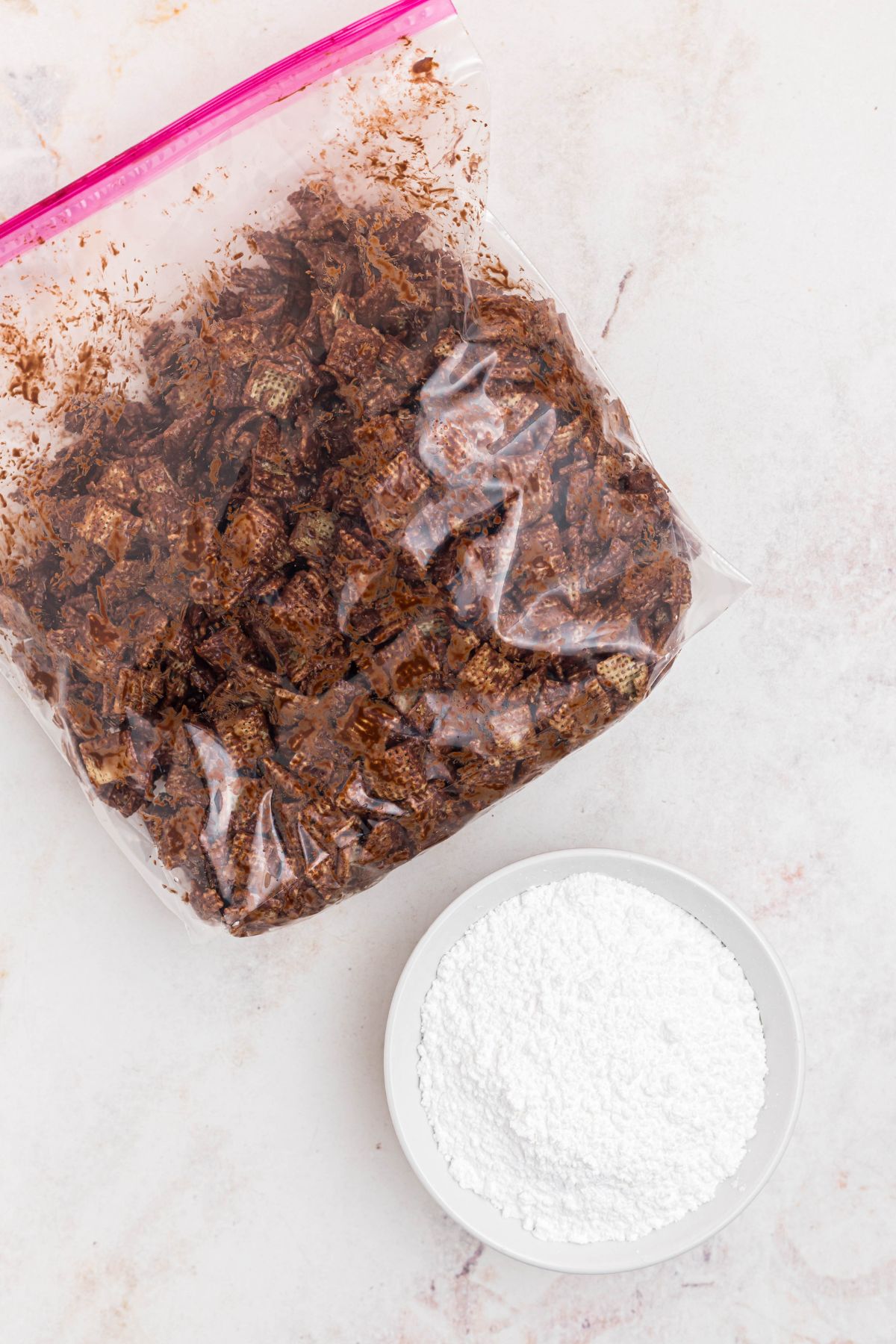 Chocolate coated cereal in a large bag, with a bowl of powdered sugar next to the bag, before mixing together. 