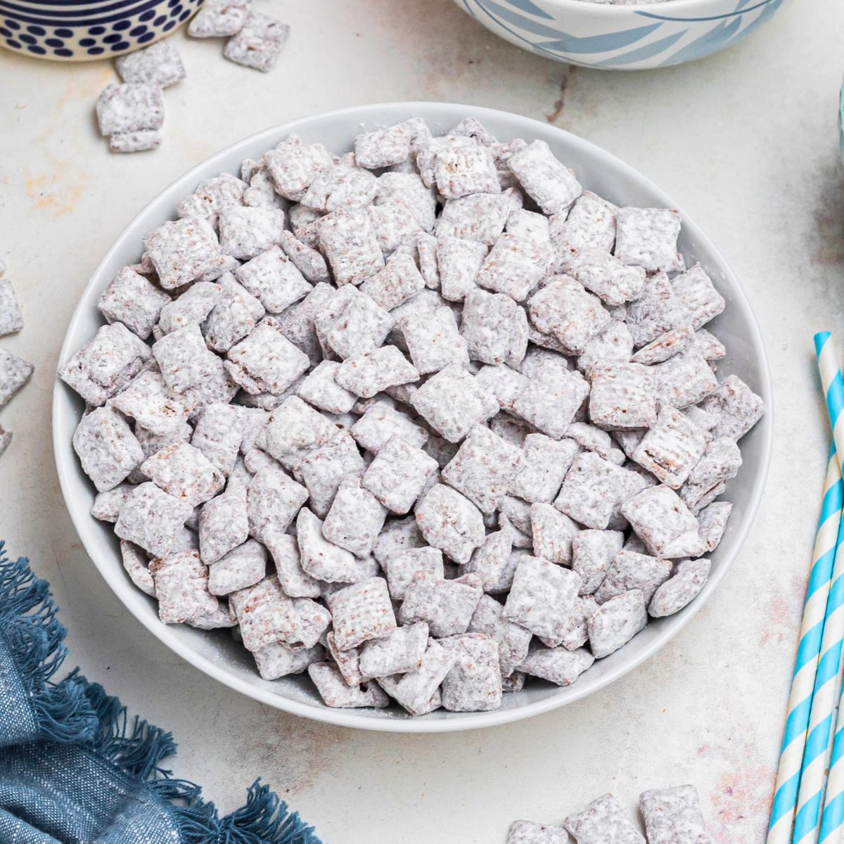 White bowl filled with powdered sugar coated cereal pieces, known as Puppy Chow, with blue linen and straws on the table.