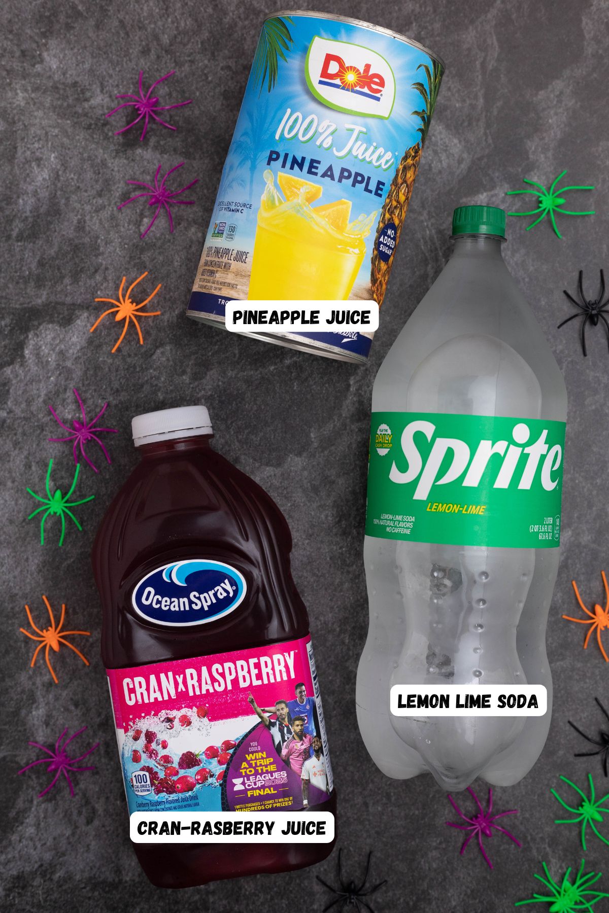 Bottles of pineapple juice, cran-raspberry juice, and lemon lime soda, used to make Halloween punch, on a black marble table. 