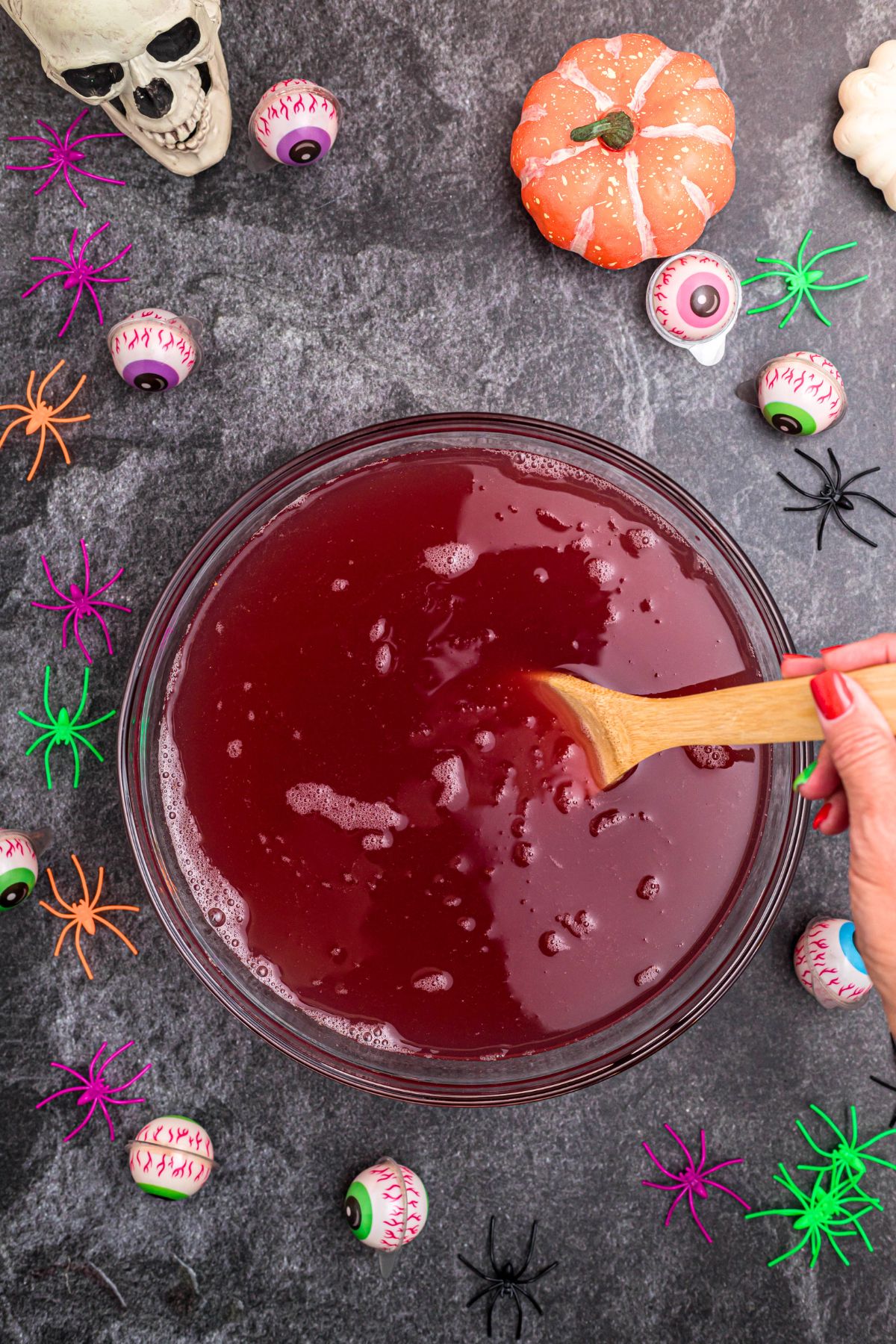 Halloween punch being stirred together with a wooden spoon with plastic spiders and candy eyes scattered around, on a black marble table. 