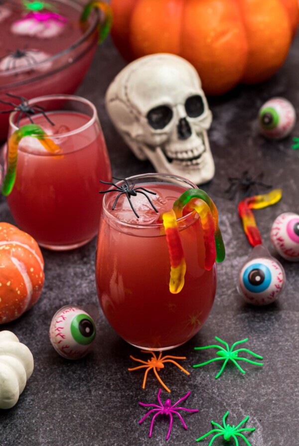 Glass of Halloween Punch in with gummy worms hanging over the side of the glass, in front of plastic skull and spiders.