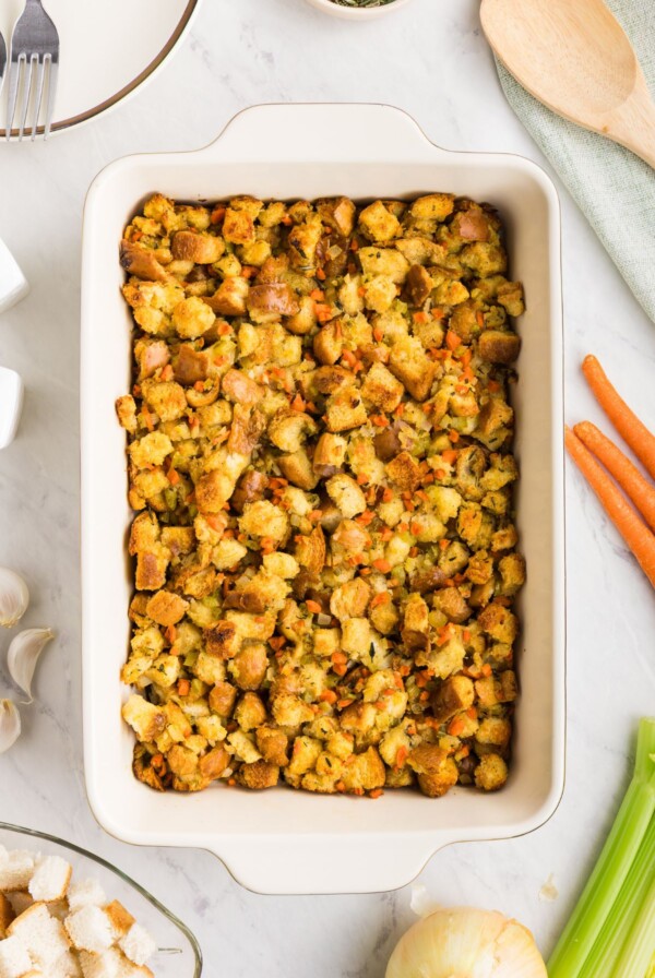 Golden, juicy stuffing in a rectangle container on a table with carrots, celery, and other ingredients around the dish.