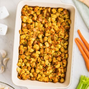 Golden, juicy stuffing in a rectangle container on a table with carrots, celery, and other ingredients around the dish.