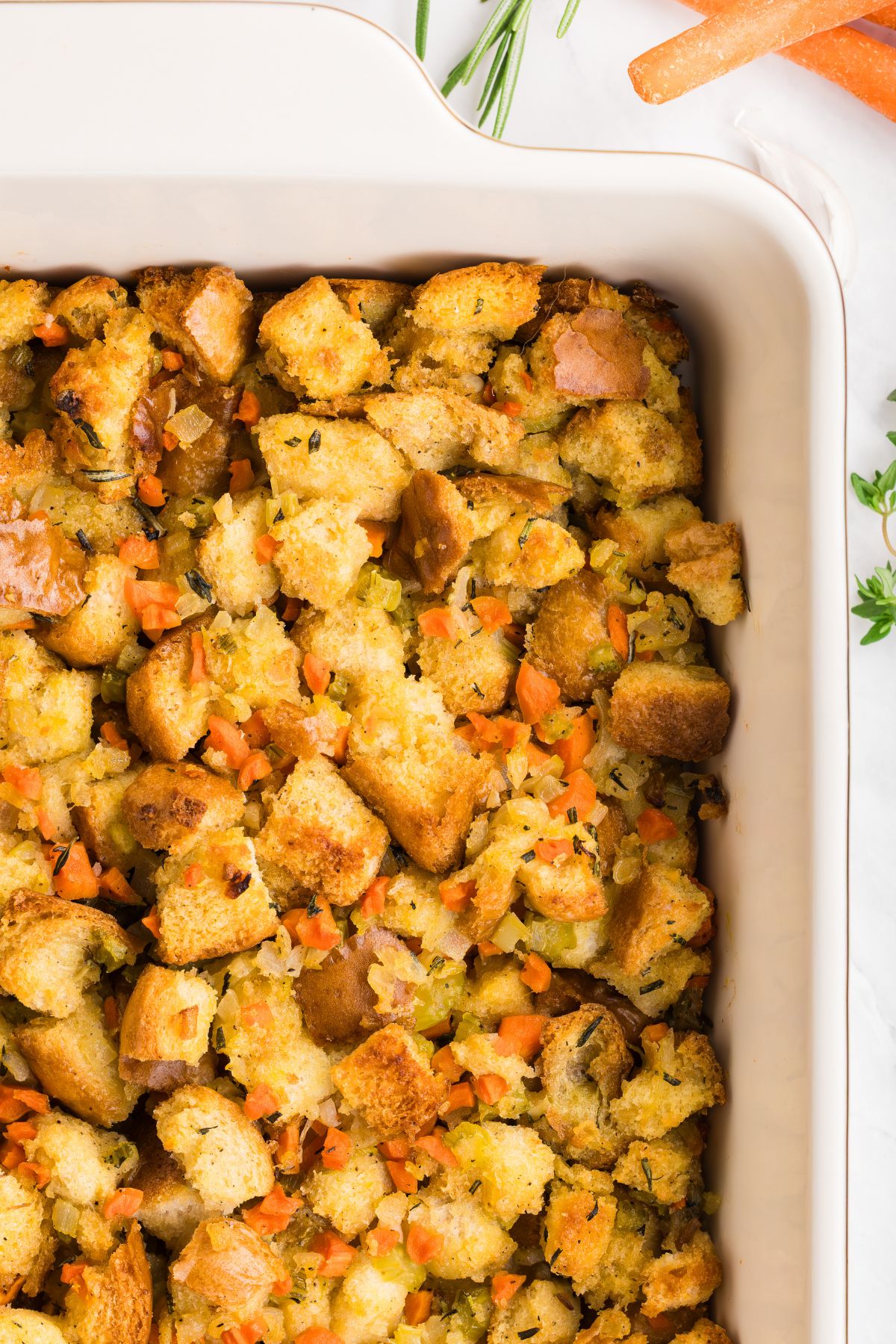 Golden, juicy stuffing in a rectangle container on a table with carrots, celery, and other ingredients around the dish.