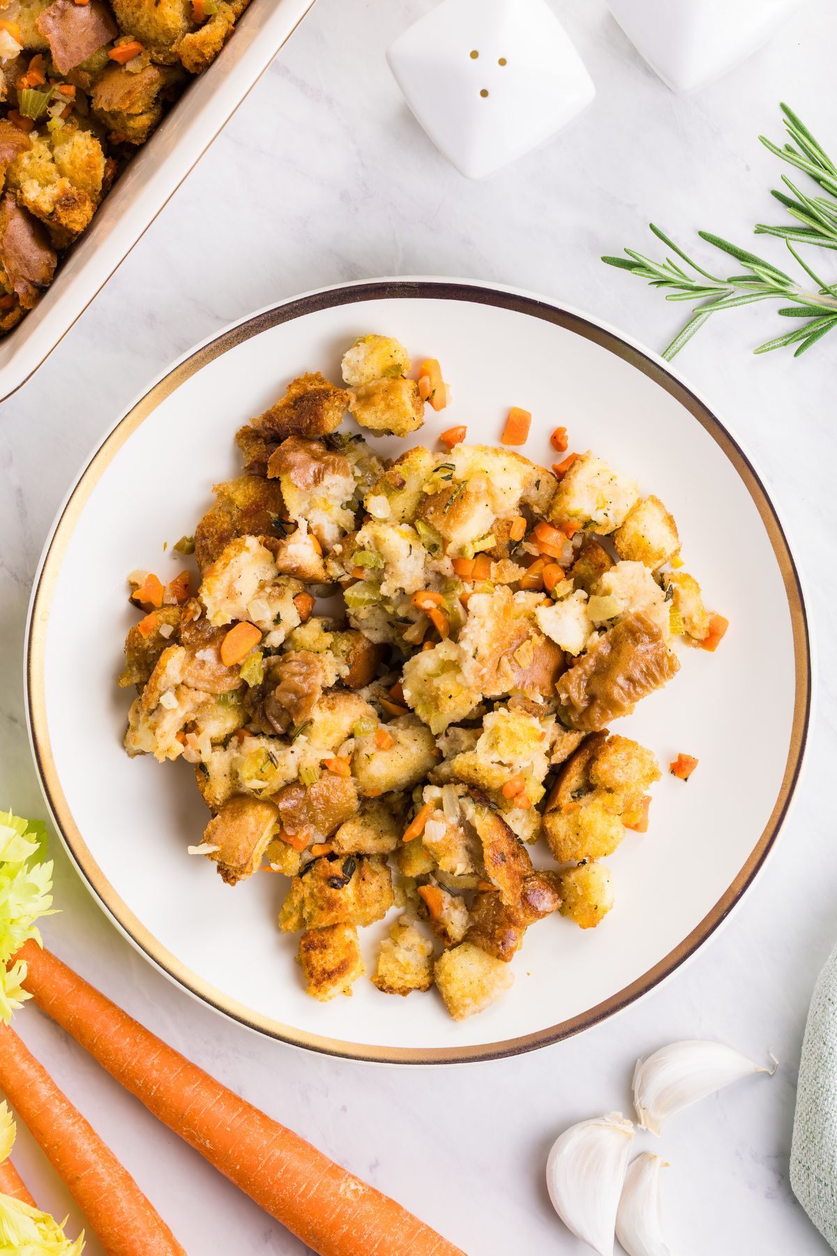 Juicy, golden brown stuffing, served on a white plate, with carrots, garlic, and celery on the table.