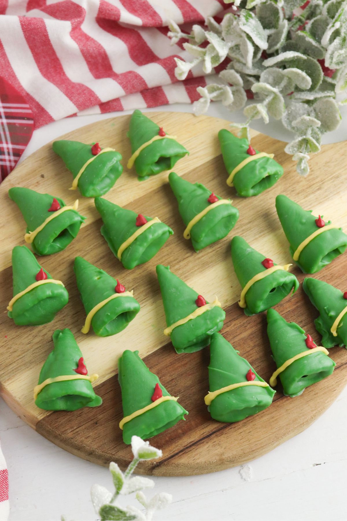 Chocolate coated green bugle chips, decorated to look like elf hats with  yellow band and red feather, on a round wooden cutting board. 