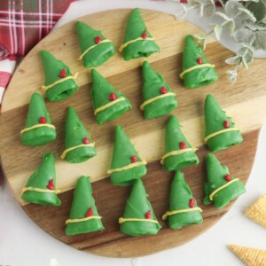 Chocolate coated green bugle chips, decorated to look like elf hats with yellow band and red feather, on a round wooden cutting board.