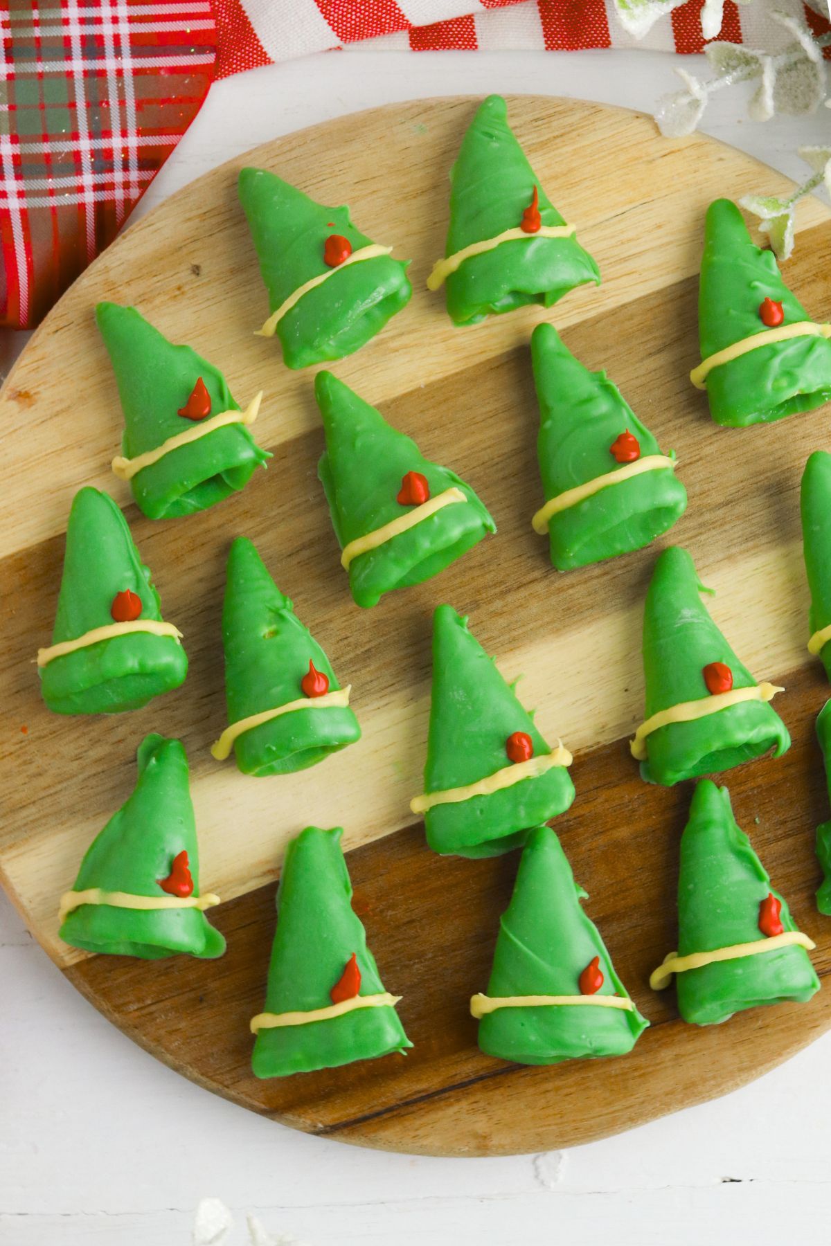 Chocolate coated green bugle chips, decorated to look like elf hats with  yellow band and red feather, on a round wooden cutting board. 