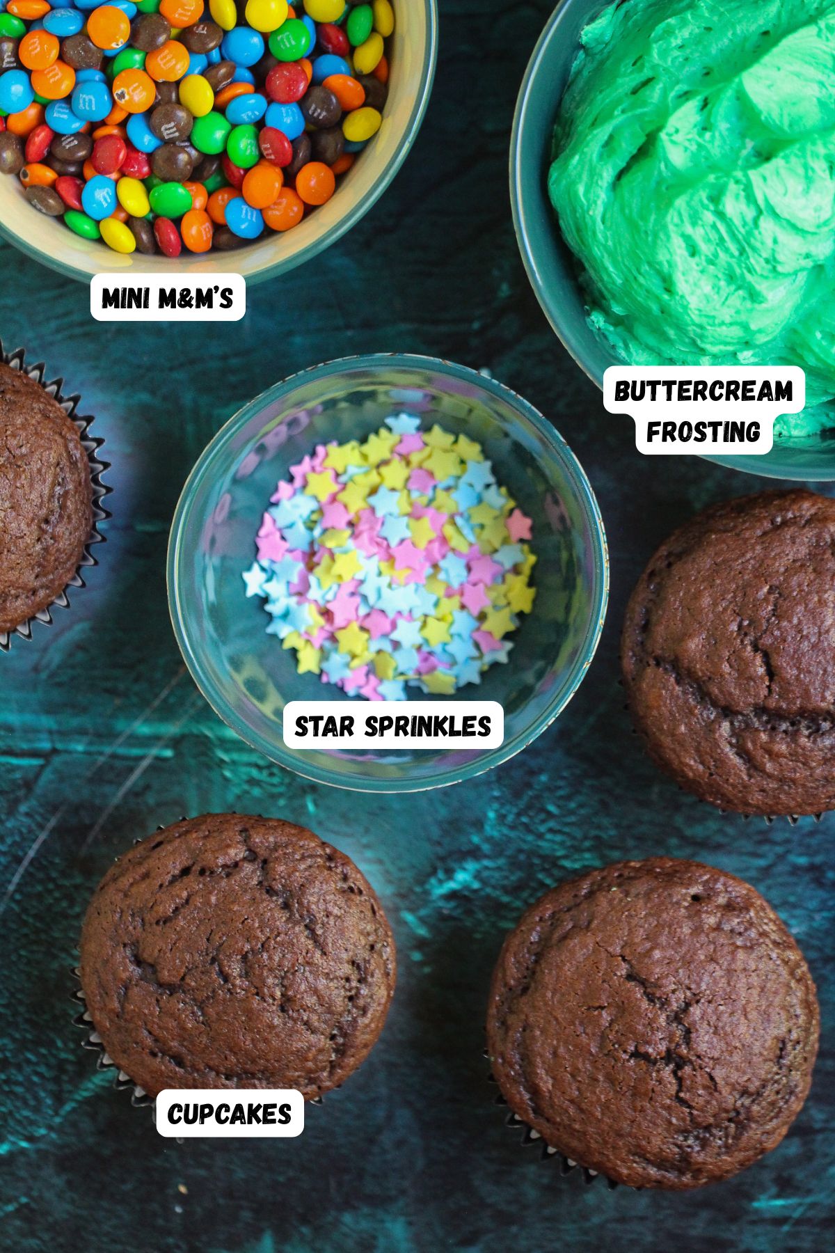 Ingredients of cupcakes, sprinkles, and frosting on a marble table, and labeled in photo.