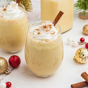 Creamy eggnog in a clear glass, topped with whipped cream and a cinnamon stick.