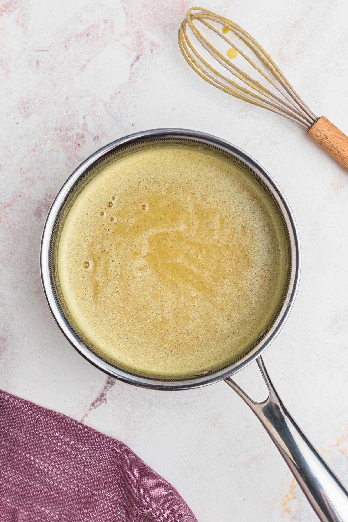 Cooked eggnog in a saucepan, with a whisk on the table next to the saucepan.