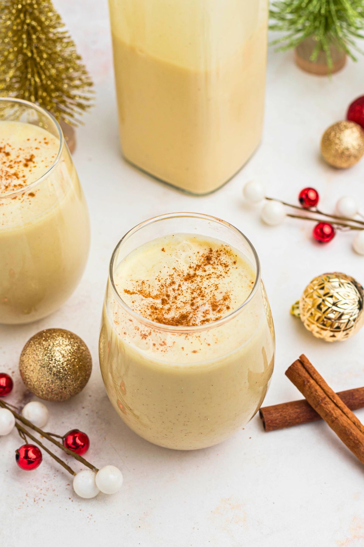 Creamy homemade eggnog in a clear glass bowl surrounded by cinnamon sticks and small Christmas decorations.