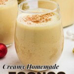 Creamy homemade eggnog in a clear glass bowl surrounded by cinnamon sticks and small Christmas decorations.