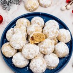 Powder sugar coated Snowball cookies, stacked on a blue plate with holiday party favors on the table around the plate, with one cookie missing a bite.